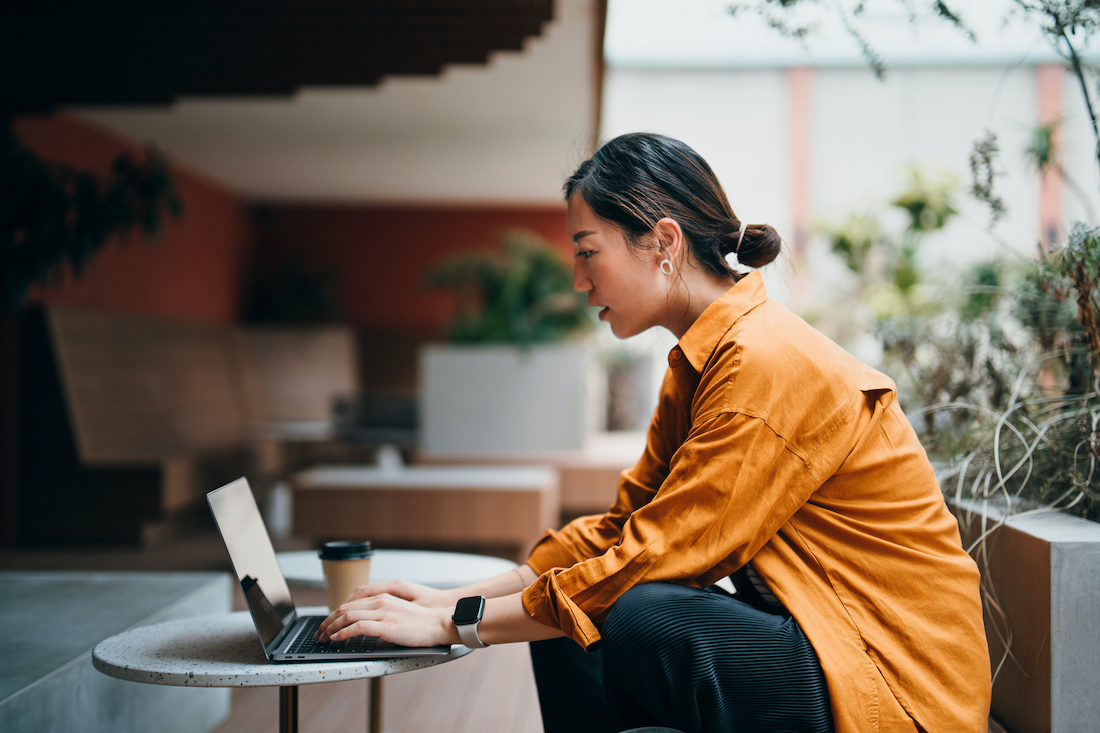 Confident young businesswoman working on laptop with business clients in outdoors co-working space, surrounded by green plants. 