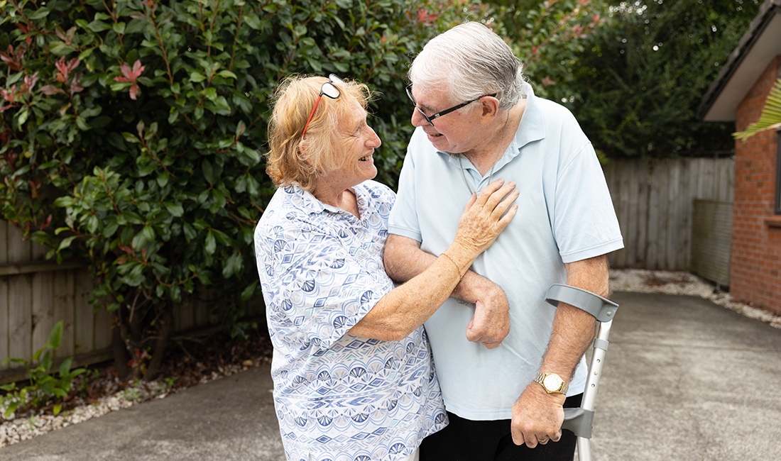 Smiling Senior Man With Dementia Standing Outside With Crutch Looking At Smiling Senior Woman Holding Him Close