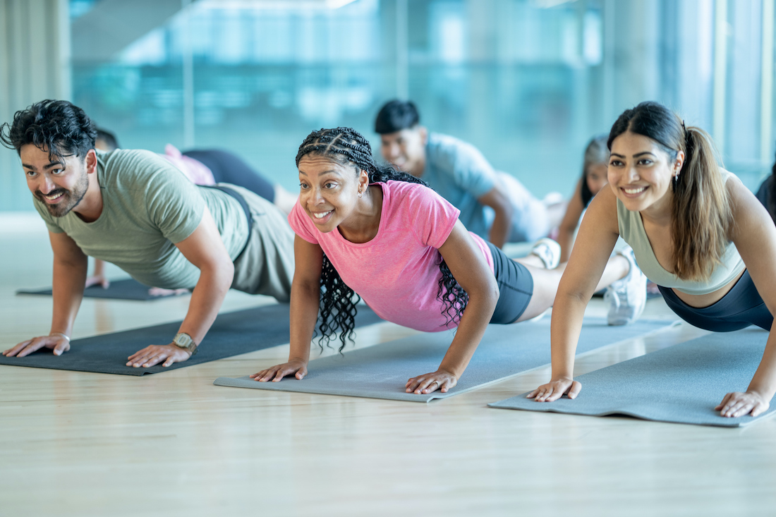 A small group of mature adults lay out spread apart on yoga mats in a gym, as they participate in a fitness class together.