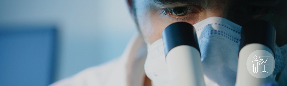 masked researcher looks thru lens of microscope