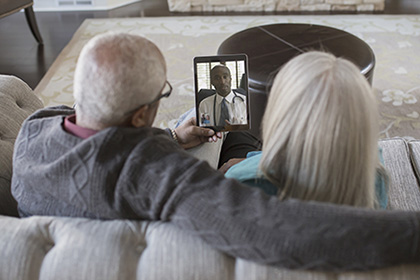 A man and a woman sit on a couch and talk to a doctor on a tablet